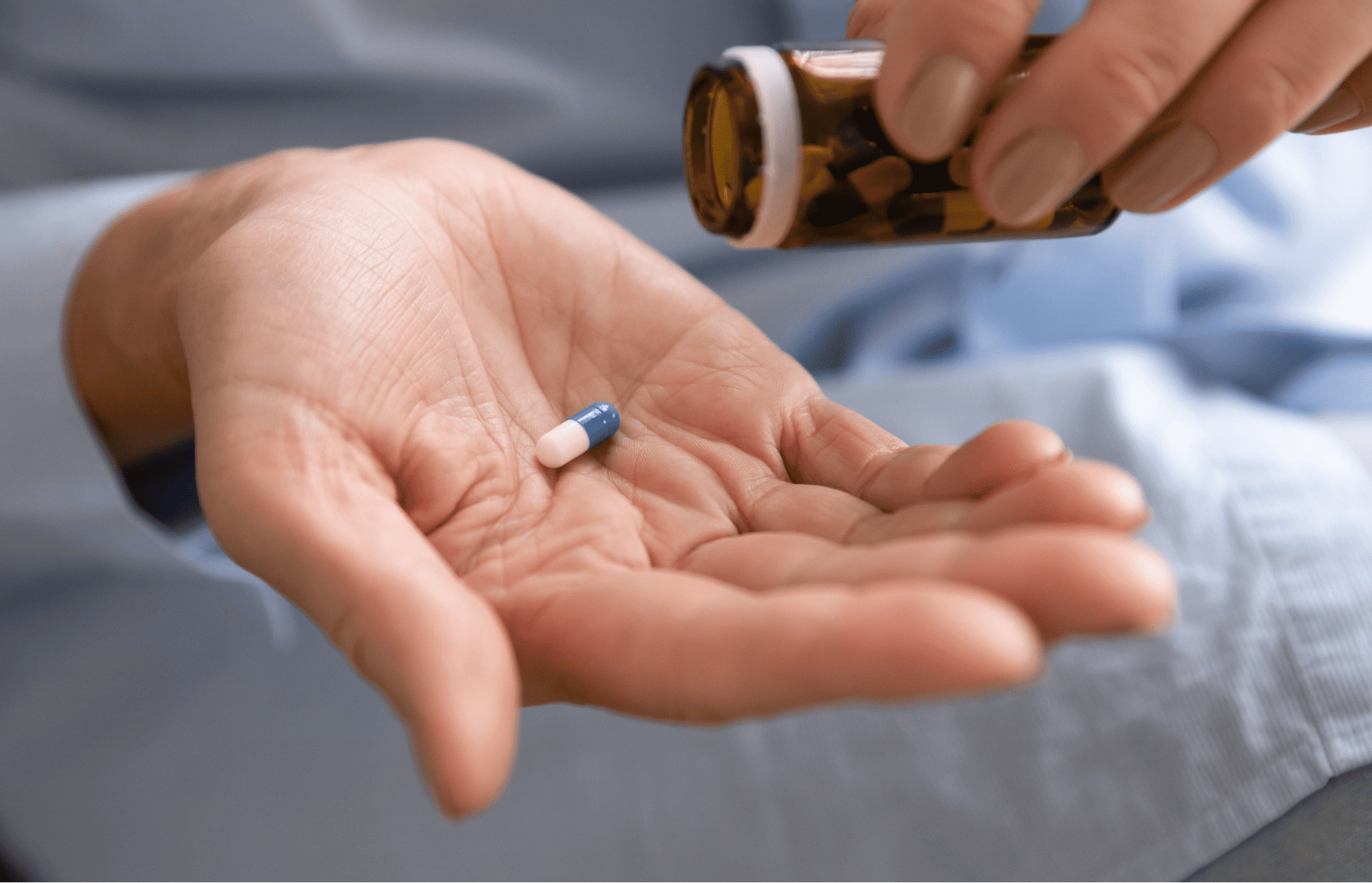 A hand holds a blue and white capsule pill while someone pours medication from a brown bottle above it